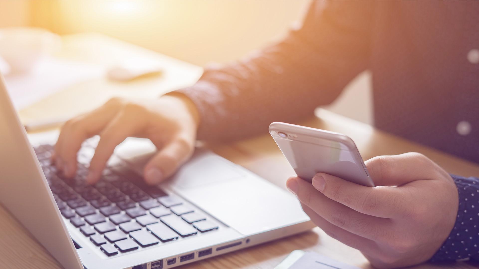 Person hält ein Smartphone in der Hand und tippt auf einem Laptop.